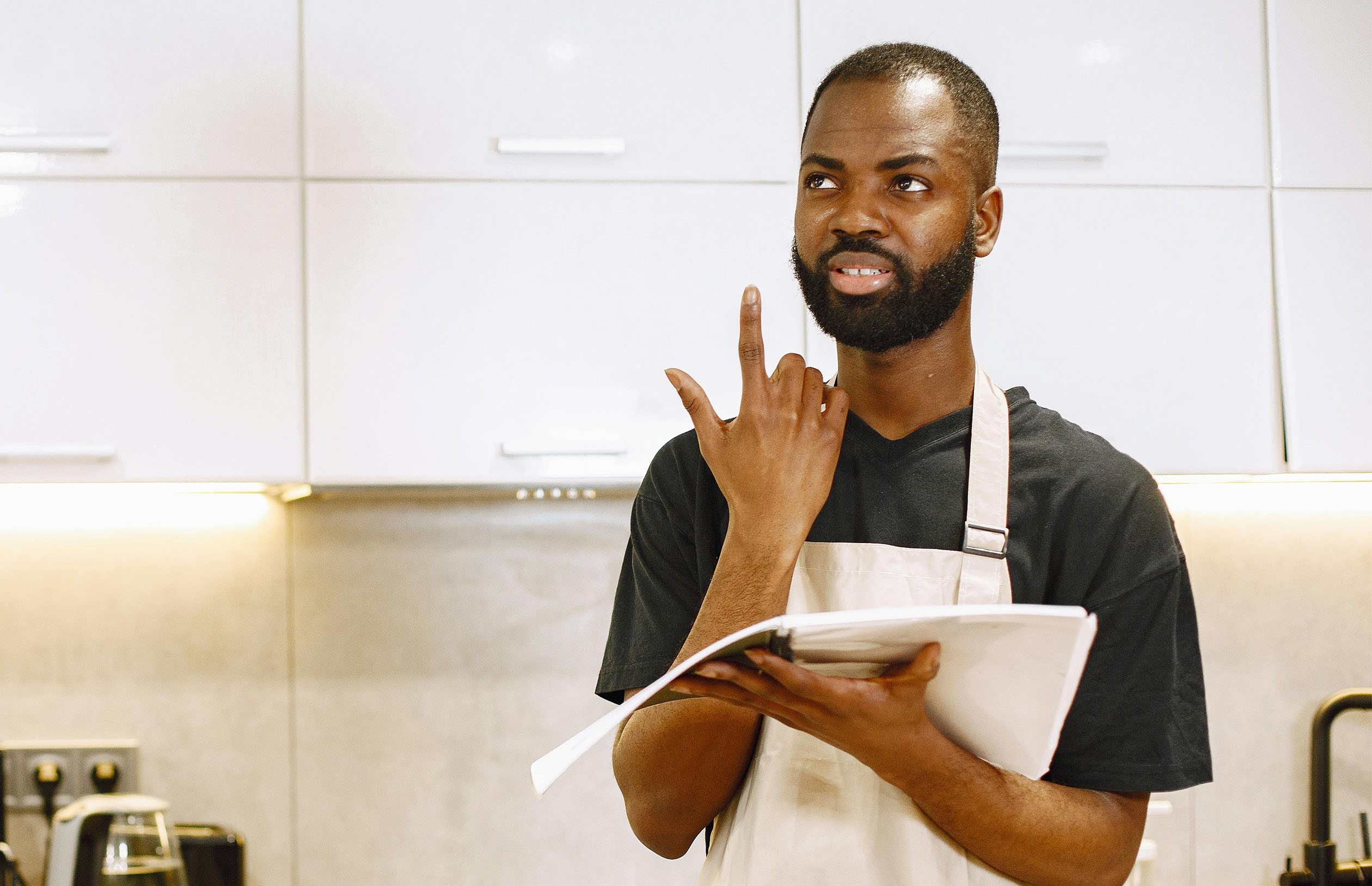 A man in a black shirt and beige apron holding a notebook and thinking in a kitchen.