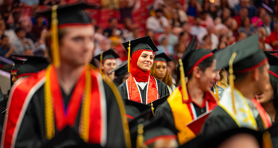 A graduation ceremony at the College of Liberal Arts and Social Sciences showing graduates in black caps and gowns with colorful academic regalia, with a smiling female graduate wearing a red hijab prominently featured in the center. The scene captures a formal commencement event with graduates holding their caps and attendees visible in stadium seating in the background.